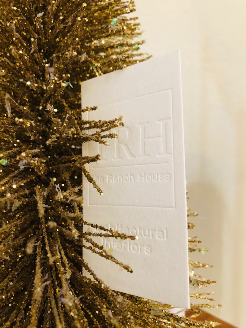 以下が含まれることがあります： TRH The Ranch House Architectural Interiorsとエンボス加工された白い長方形の名刺。光を反射する細い針のような枝を持つ、きらめく金色の人工の木の隣に配置されています。