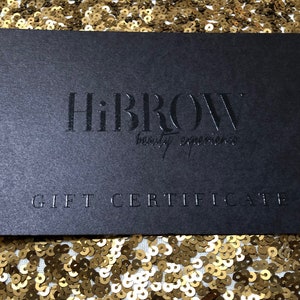 May include: A rectangular black gift certificate featuring the embossed text HiBROW beauty experience in the center and the words GIFT CERTIFICATE printed in capital letters at the bottom, presented on a surface covered in gold sequins.