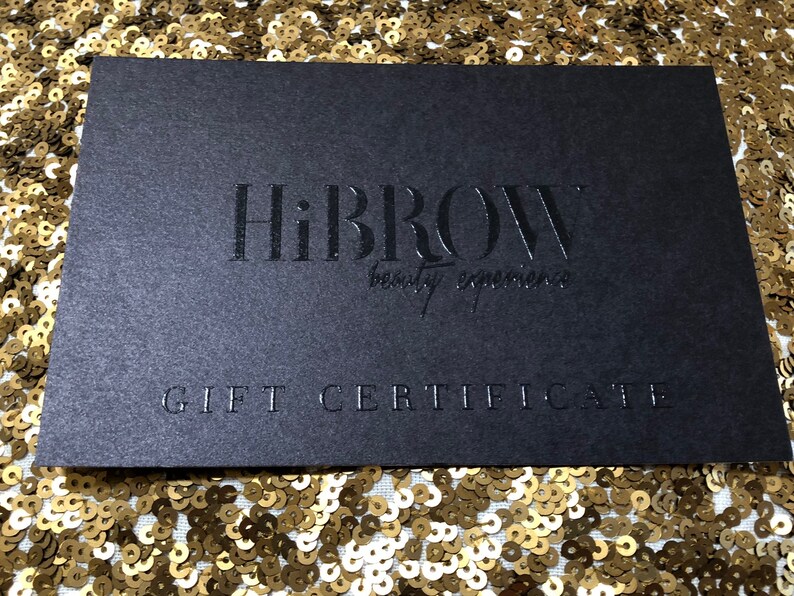 May include: A rectangular black gift certificate featuring the embossed text HiBROW beauty experience in the center and the words GIFT CERTIFICATE printed in capital letters at the bottom, presented on a surface covered in gold sequins.