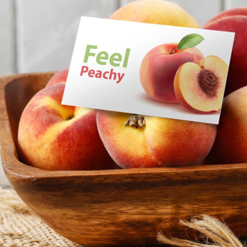 K&ouml;nnte beinhalten: Eine Holzschale gef&uuml;llt mit reifen Pfirsichen. Die Pfirsiche sind eine Mischung aus Rot und Gelb mit einer flauschigen Haut. Eine Karte mit dem Text "Feel Peachy" liegt auf den Pfirsichen.