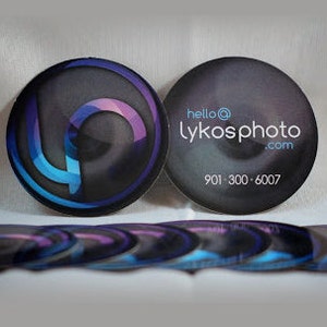 Puede incluir: Una pila de tarjetas de visita circulares con un fondo oscuro, un logotipo abstracto azul y morado en un lado y el texto hello@lykosphoto.com con el n&uacute;mero de tel&eacute;fono 901-300-6007 impreso en blanco en el otro lado de las tarjetas.