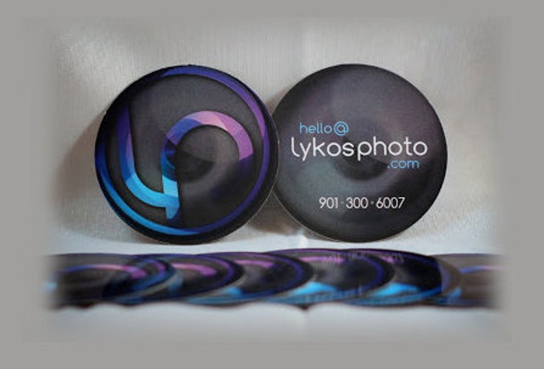 Puede incluir: Una pila de tarjetas de visita circulares con un fondo oscuro, un logotipo abstracto azul y morado en un lado y el texto hello@lykosphoto.com con el n&uacute;mero de tel&eacute;fono 901-300-6007 impreso en blanco en el otro lado de las tarjetas.