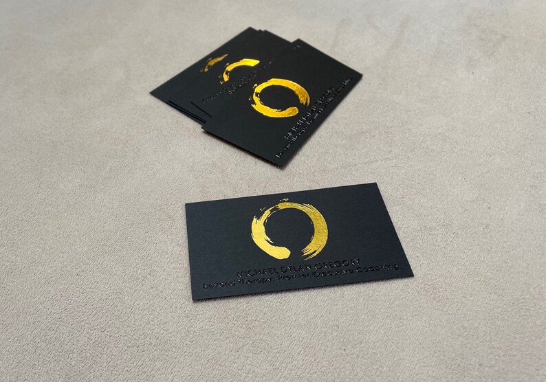 May include: A stack of rectangular black business cards and one single card placed in front. Each card features a gold foil-stamped circular brushstroke logo, with the text MICHAEL DYLAN GREGORY and BEYOND THERAPY: PREMIER EXECUTIVE COACHING printed below.