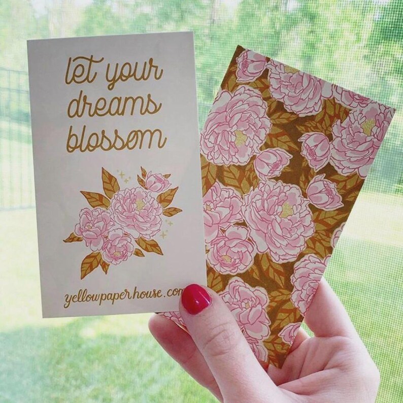 May include: Two rectangular paper cards. The left card is white with the gold text "let your dreams blossom" above a cluster of pink peonies and gold leaves, with "yellowpaperhouse.com" at the bottom. The right card features a floral pattern.