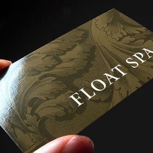 May include: A rectangular business card with a glossy finish, featuring a dark olive green background with an intricate, tone-on-tone botanical leaf pattern. The text FLOAT SPA is printed in a clean, white, sans-serif font on the right side.