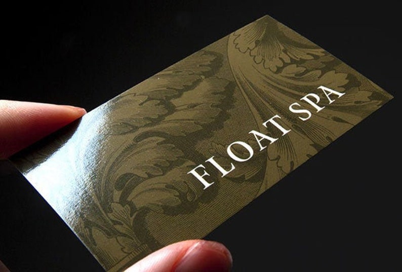May include: A rectangular business card with a glossy finish, featuring a dark olive green background with an intricate, tone-on-tone botanical leaf pattern. The text FLOAT SPA is printed in a clean, white, sans-serif font on the right side.