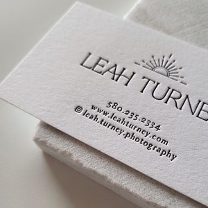 以下が含まれることがあります： 質感のある白い名刺。エンボス加工された「LEAH TURNER」の文字、太陽のアイコン、電話番号「580.235.2334」、ウェブサイト「www.leahturney.com」、Instagramアカウント「leah.turney.photography」がダークグレーのインクで印刷されています。
