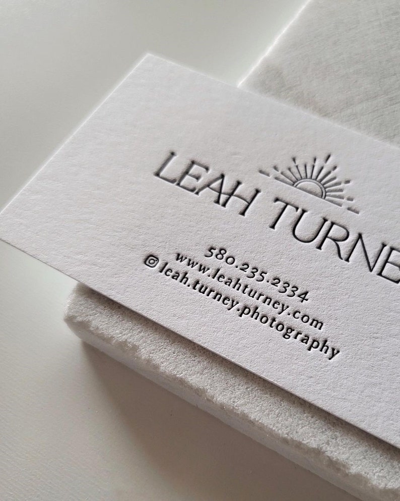 以下が含まれることがあります： 質感のある白い名刺。エンボス加工された「LEAH TURNER」の文字、太陽のアイコン、電話番号「580.235.2334」、ウェブサイト「www.leahturney.com」、Instagramアカウント「leah.turney.photography」がダークグレーのインクで印刷されています。