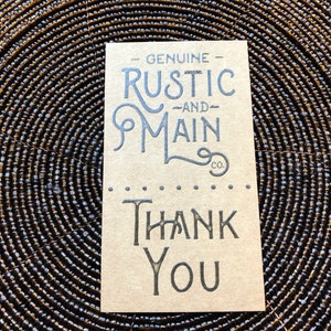 May include: A rectangular tan card with dark grey printed text. The top portion reads GENUINE RUSTIC AND MAIN CO. in a stylised font. Below a horizontal line of dots, the bottom portion features the words THANK YOU in a bold, serif typeface.