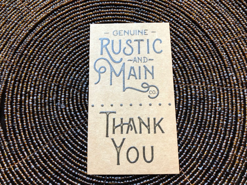 May include: A rectangular tan card with dark grey printed text. The top portion reads GENUINE RUSTIC AND MAIN CO. in a stylised font. Below a horizontal line of dots, the bottom portion features the words THANK YOU in a bold, serif typeface.