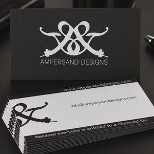 May include: A stack of rectangular business cards with a black and white color scheme. The top card is black with a white logo and the text AMPERSAND DESIGNS. The lower cards are white with a black logo, website, email, and the text because everyone is entitled to a charmed life.