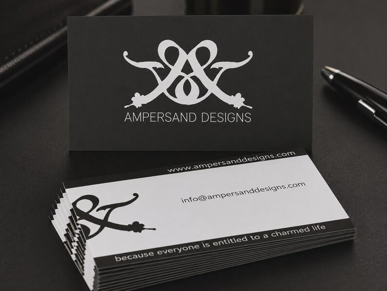 May include: A stack of rectangular business cards with a black and white color scheme. The top card is black with a white logo and the text AMPERSAND DESIGNS. The lower cards are white with a black logo, website, email, and the text because everyone is entitled to a charmed life.