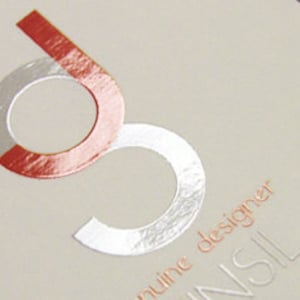 May include: A light beige card featuring a stylized logo of the letter g in a metallic silver and orange foil finish. Below the logo, the text genuine designer is printed in a small orange font, with the letters NSIL visible in a larger embossed style.