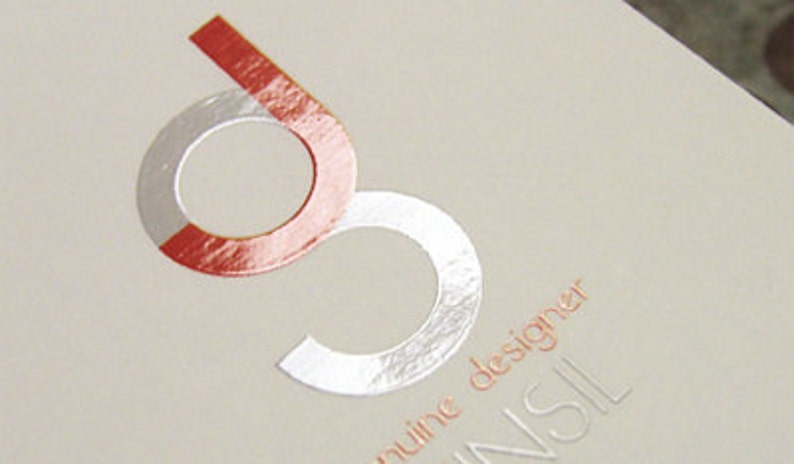 May include: A light beige card featuring a stylized logo of the letter g in a metallic silver and orange foil finish. Below the logo, the text genuine designer is printed in a small orange font, with the letters NSIL visible in a larger embossed style.