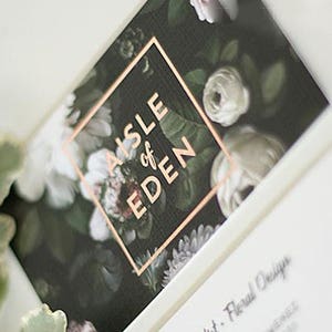 May include: A black and white floral design with a copper square overlay that says "AISLE of EDEN".