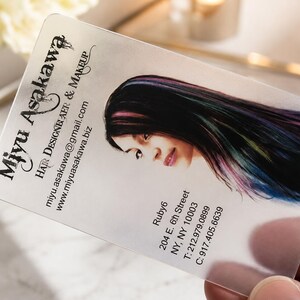 May include: A translucent rectangular business card featuring the text Miyu Asakawa, Hair Designer & Makeup, an email address, a website, and an address at 204 E. 6th Street, NY, NY 10003, alongside a portrait of a person with dark, multi-colored hair.