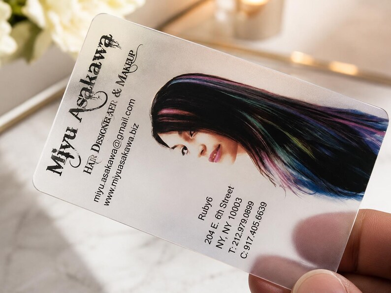 May include: A translucent rectangular business card featuring the text Miyu Asakawa, Hair Designer & Makeup, an email address, a website, and an address at 204 E. 6th Street, NY, NY 10003, alongside a portrait of a person with dark, multi-colored hair.