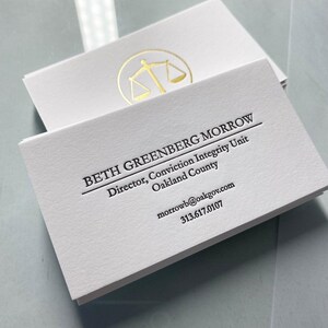 May include: A stack of white business cards featuring black text that reads BETH GREENBERG MORROW, Director, Conviction Integrity Unit, Oakland County, morrowb@oakgov.com, and 313.617.0107. A gold foil scale of justice icon is printed on the top card.