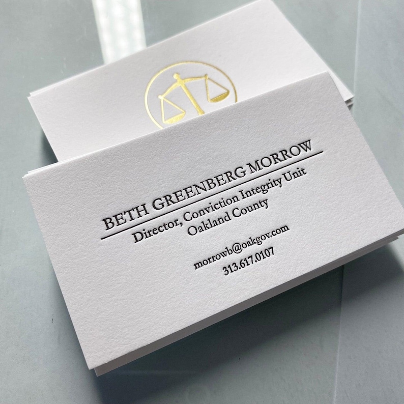 May include: A stack of white business cards featuring black text that reads BETH GREENBERG MORROW, Director, Conviction Integrity Unit, Oakland County, morrowb@oakgov.com, and 313.617.0107. A gold foil scale of justice icon is printed on the top card.