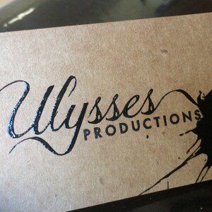 May include: A business card with the name "Ulysses Productions" in elegant script and sans-serif font. The card is a light brown color with a black ink blotch design.