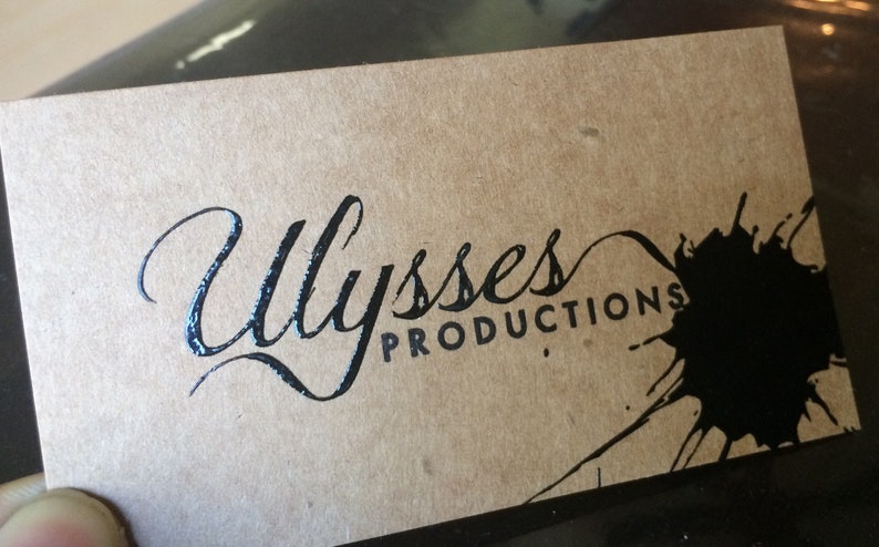 May include: A business card with the name "Ulysses Productions" in elegant script and sans-serif font. The card is a light brown color with a black ink blotch design.