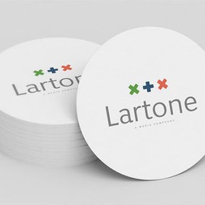Puede incluir: Una pila de posavasos circulares blancos con el logotipo de Lartone en texto gris y un s&iacute;mbolo de cruz verde, azul oscuro y rojo encima. El texto debajo del logo dice A MEDIA COMPANY en letras may&uacute;sculas peque&ntilde;as de color gris.