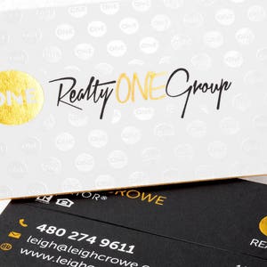 May include: A white business card with a gold foil circle logo and the text Realty ONE Group, featuring a repeating pattern of the word ONE. Below it, a black business card displays gold foil text, a phone number, email, and website address.