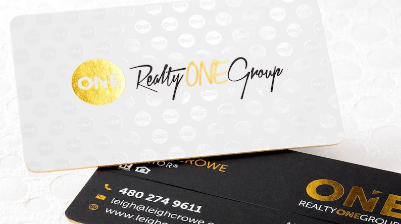 May include: A white business card with a gold foil circle logo and the text Realty ONE Group, featuring a repeating pattern of the word ONE. Below it, a black business card displays gold foil text, a phone number, email, and website address.