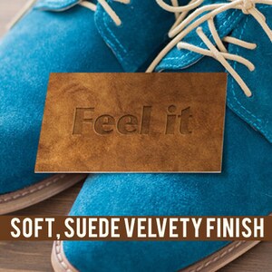 K&ouml;nnte beinhalten: Nahaufnahme eines Paares blauer Wildlederschuhe mit wei&szlig;en Schn&uuml;rsenkeln. Ein braunes Lederst&uuml;ck mit dem Text "Feel it" liegt auf den Schuhen. Der Text "SOFT, SUEDE VELVETY FINISH" wird unter den Schuhen angezeigt.