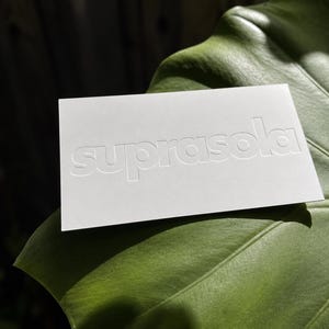 May include: A rectangular white card with the word suprasola embossed in lowercase letters across the center. The card features a clean, matte finish and is positioned horizontally, displaying a crisp, tactile impression of the brand name.
