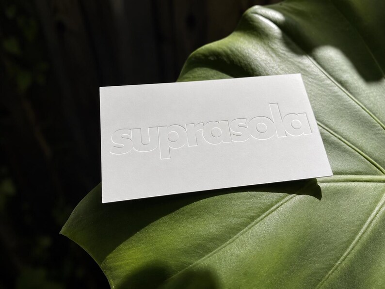 May include: A rectangular white card with the word suprasola embossed in lowercase letters across the center. The card features a clean, matte finish and is positioned horizontally, displaying a crisp, tactile impression of the brand name.