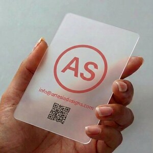 May include: A rectangular, semi-transparent frosted business card featuring a coral-colored circular logo with the letters AS inside. Below the logo, the text info@artesiadesigns.com is printed in coral, followed by a black square QR code.