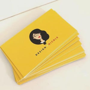 May include: A stack of rectangular business cards in a bright yellow color. Each card features a black graphic illustration of a person with dark hair and red lips, accompanied by the black text "DESIGN x NICOLE" printed below the illustration.