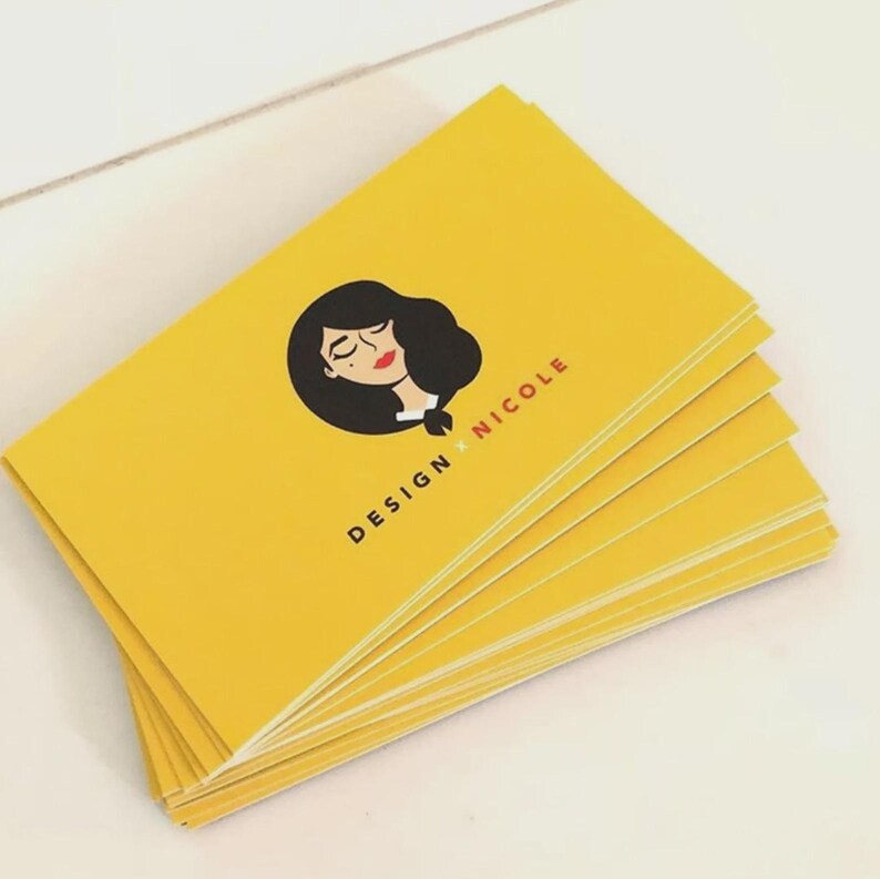 May include: A stack of rectangular business cards in a bright yellow color. Each card features a black graphic illustration of a person with dark hair and red lips, accompanied by the black text "DESIGN x NICOLE" printed below the illustration.