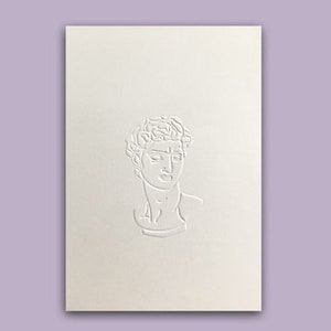 May include: A cream-colored card with a raised relief of a classical bust. The bust is centered on the card and is a detailed depiction of a male figure with curly hair and a serene expression. The background is a soft lavender.