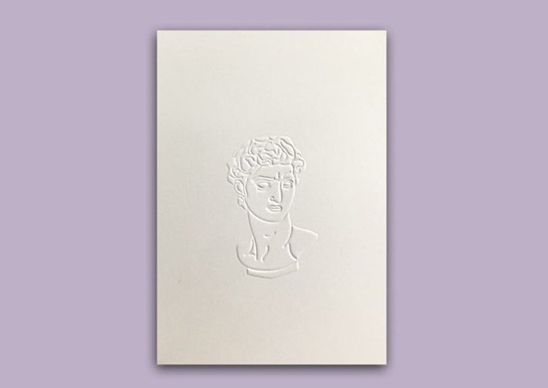 May include: A cream-colored card with a raised relief of a classical bust. The bust is centered on the card and is a detailed depiction of a male figure with curly hair and a serene expression. The background is a soft lavender.