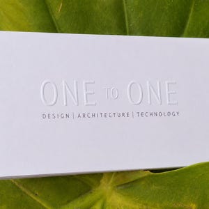 May include: A rectangular white business card with embossed text reading ONE TO ONE in the center. Below this, the words DESIGN | ARCHITECTURE | TECHNOLOGY are printed in a smaller, dark gray sans-serif font. The card has a clean, matte finish.