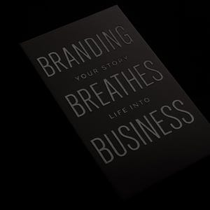 May include: A rectangular black card featuring embossed silver lettering that reads BRANDING YOUR STORY BREATHES LIFE INTO BUSINESS. The text is arranged in a multi-line layout with a clean, sans-serif font style against a solid dark background.