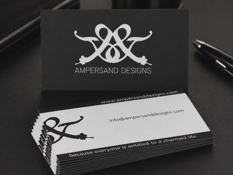 May include: A stack of rectangular business cards with a black and white color scheme. The top card is black with a white logo and the text AMPERSAND DESIGNS. The lower cards are white with a black logo, website, email, and the text because everyone is entitled to a charmed life.