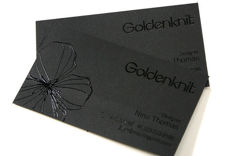 May include: Two rectangular black business cards featuring the text Goldenknit in a raised glossy finish. The cards display a glossy embossed floral graphic on the left side and include the name Nina Thomas with contact details printed in black.