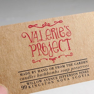 May include: A rectangular tan business card with a textured paper finish. The text VALERIE'S PROJECT is printed in red ink with decorative flourishes. Below, black text reads MADE BY HAND OR FROM THE GARDEN, candles bathbombs soap preserves, 99 WILLOW AVENUE JEFFERSON PINES, KINGSTON NOVA SCOTIA.