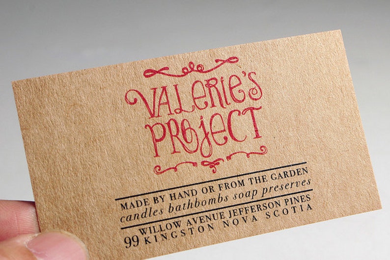 May include: A rectangular tan business card with a textured paper finish. The text VALERIE'S PROJECT is printed in red ink with decorative flourishes. Below, black text reads MADE BY HAND OR FROM THE GARDEN, candles bathbombs soap preserves, 99 WILLOW AVENUE JEFFERSON PINES, KINGSTON NOVA SCOTIA.