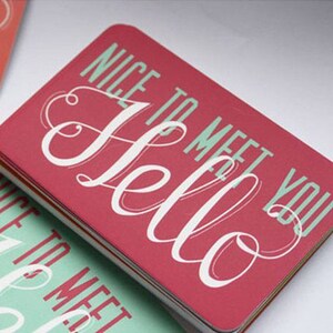 May include: A stack of rectangular business cards with rounded corners. The top card is red with the word Hello in large white script and the text NICE TO MEET YOU in light green block letters. Other cards in the stack feature inverted colors.