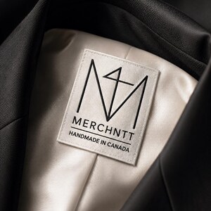 May include: A black blazer with a cream-colored satin lining. A rectangular white fabric label is sewn into the interior neck, featuring a black geometric logo, the text MERCHNTT, and the words HANDMADE IN CANADA printed in black ink.