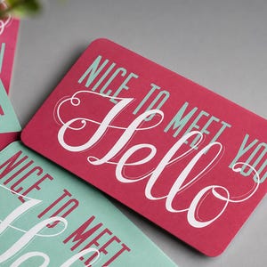May include: A rectangular card with a dark pink background featuring the white script text Hello and the light blue sans-serif text Nice to meet you. The card has rounded corners and is displayed alongside other similar cards in light blue.