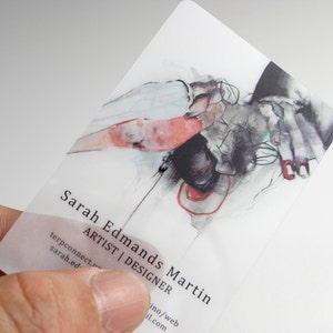 May include: A translucent rectangular business card featuring a detailed watercolor illustration of hands in shades of black, white, and red. The text reads Sarah Edmans Martin, ARTIST | DESIGNER, along with contact information in black sans-serif font.