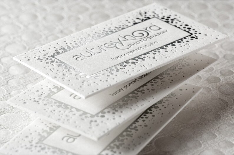May include: A stack of white rectangular business cards featuring silver foil accents in a scattered dot pattern. The top card displays the text aubrey hord photography luxury portrait studio in a clean, sans-serif font with embossed lettering.