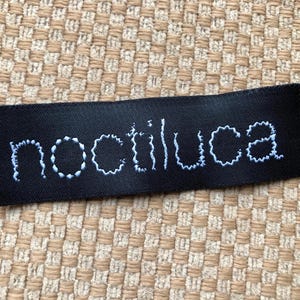 May include: A rectangular black fabric clothing label featuring the word noctiluca embroidered in white thread. The letters are rendered in a lowercase, slightly irregular, hand-stitched style font centered on the dark, smooth material surface.