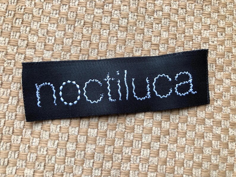May include: A rectangular black fabric clothing label featuring the word noctiluca embroidered in white thread. The letters are rendered in a lowercase, slightly irregular, hand-stitched style font centered on the dark, smooth material surface.