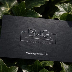 K&ouml;nnte beinhalten: Eine rechteckige schwarze Visitenkarte mit strukturierter Oberfl&auml;che und einem silbernen Pr&auml;gelogo mit der Aufschrift EMG SOLUTIONS sowie einer stilisierten Grafik aus Netzstecker und Pulslinie. Die Website www.emgsolutions.biz ist unten wei&szlig; aufgedruckt.